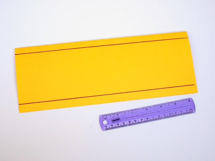 Piece of yellow construction paper folded in half with purple lines drawn on it and a ruler laying nearby