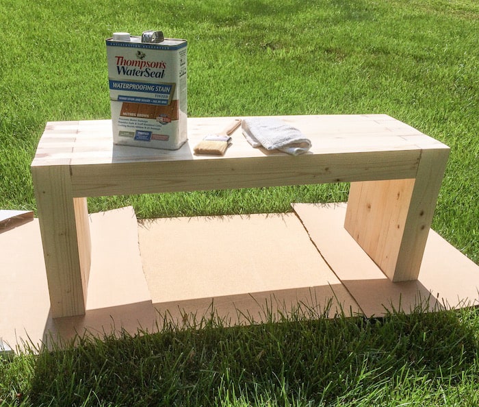 Bench sitting on cardboard with Thompson's water seal, a brush, and a rag on top