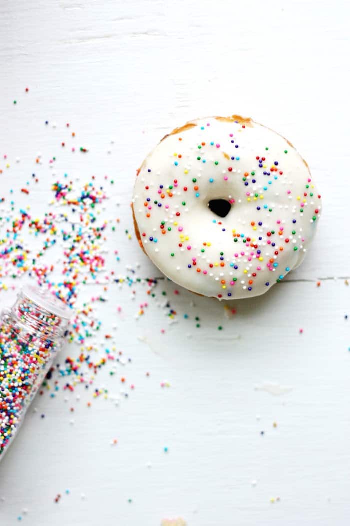 Poured out rainbow sprinkles on the table next to a baked cake donut with vanilla frosting
