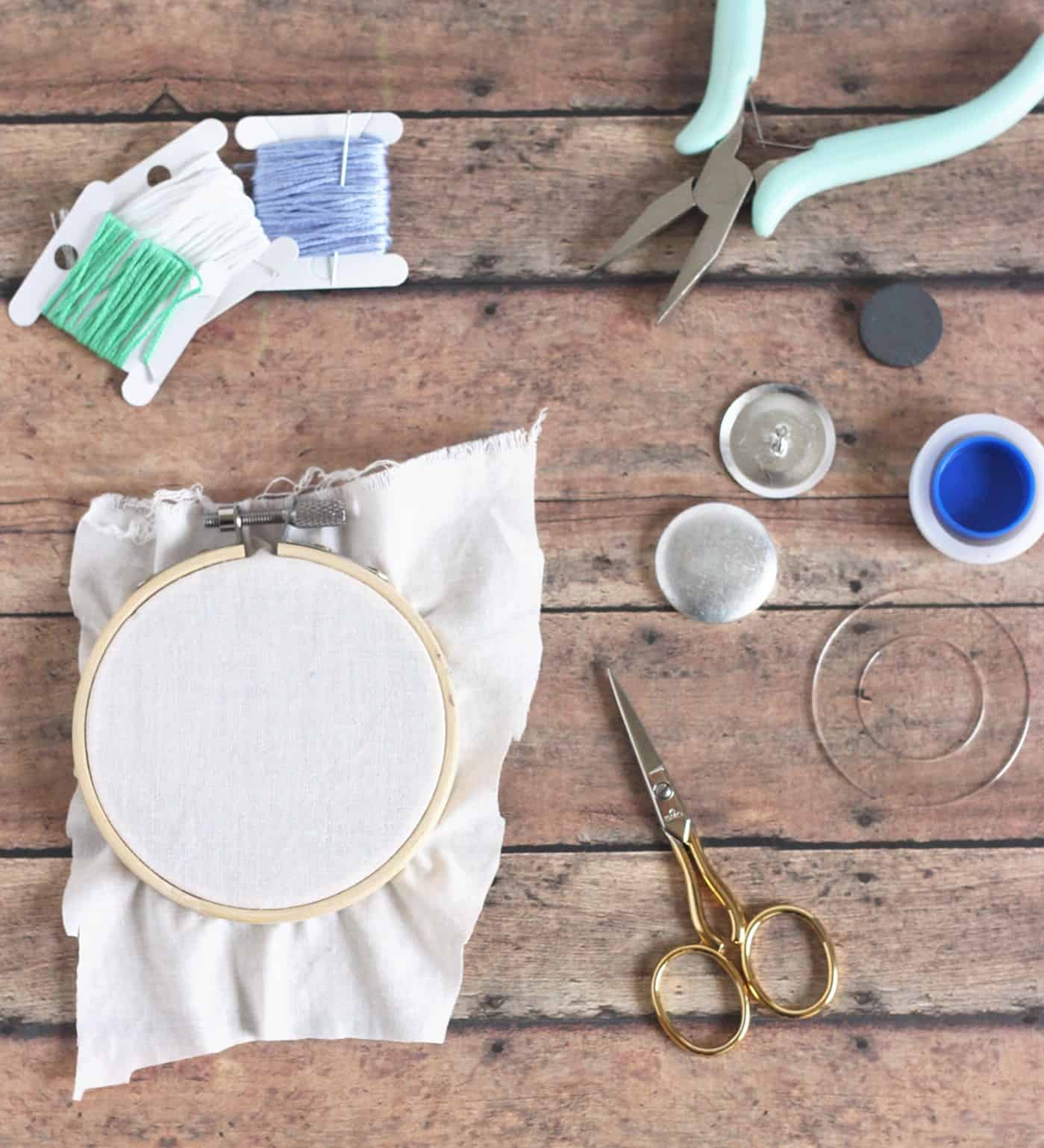 Embroidery thread, woven fabric in an embroidery hoop, jewelry pliers, and a button cover kit