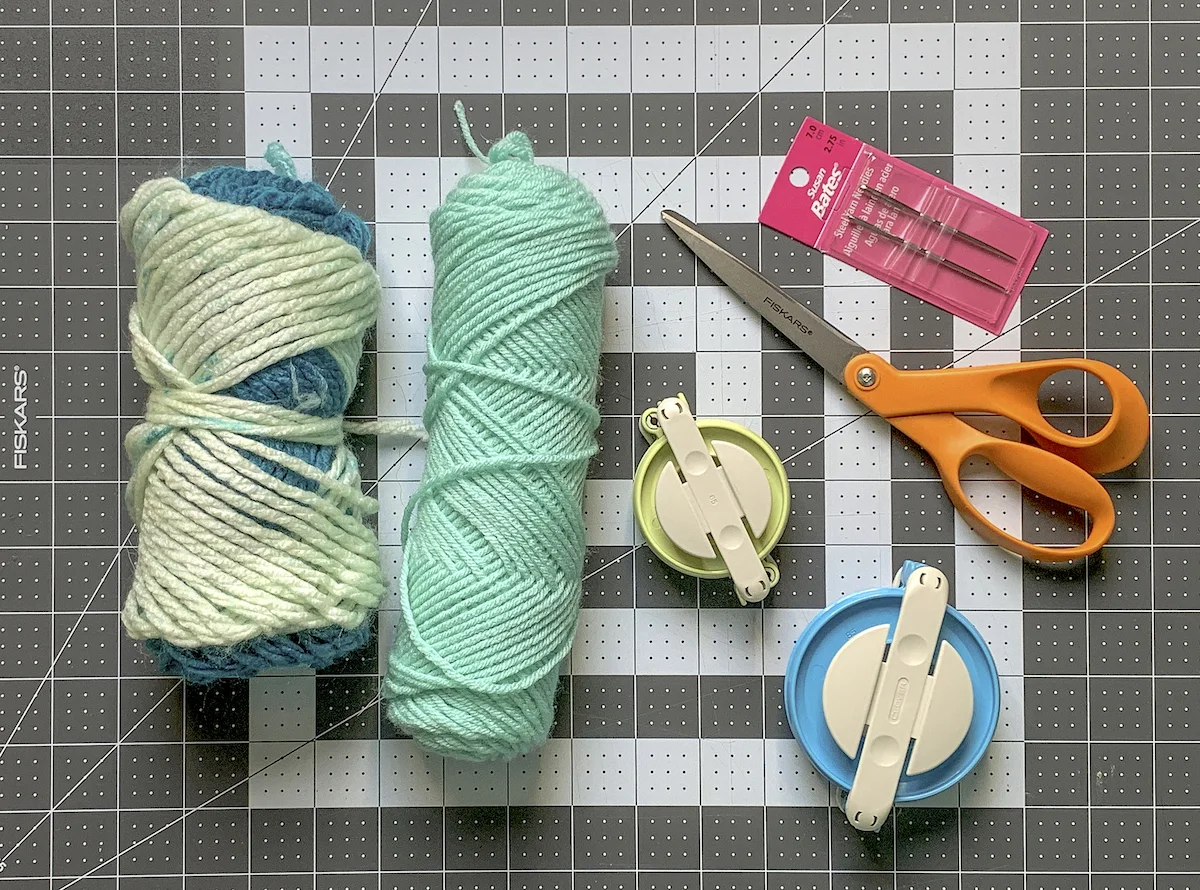 Two skeins of yarn with pom pom makers scissors and needles